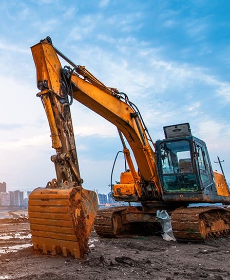 Excavadora hidráulica amarilla con orugas, operando en un sitio de construcción con suelo fangoso y edificios en el fondo. Su brazo y cuchara muestran signos de uso intenso y óxido, lo que indica un trabajo en condiciones exigentes.
