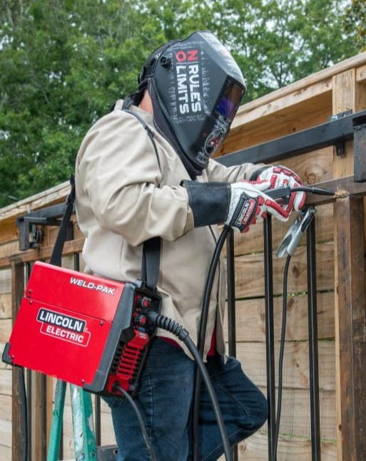 Soldador usando equipo de protección, incluyendo casco y guantes, mientras trabaja en una estructura metálica con una máquina de soldadura portátil Lincoln Electric de color rojo.